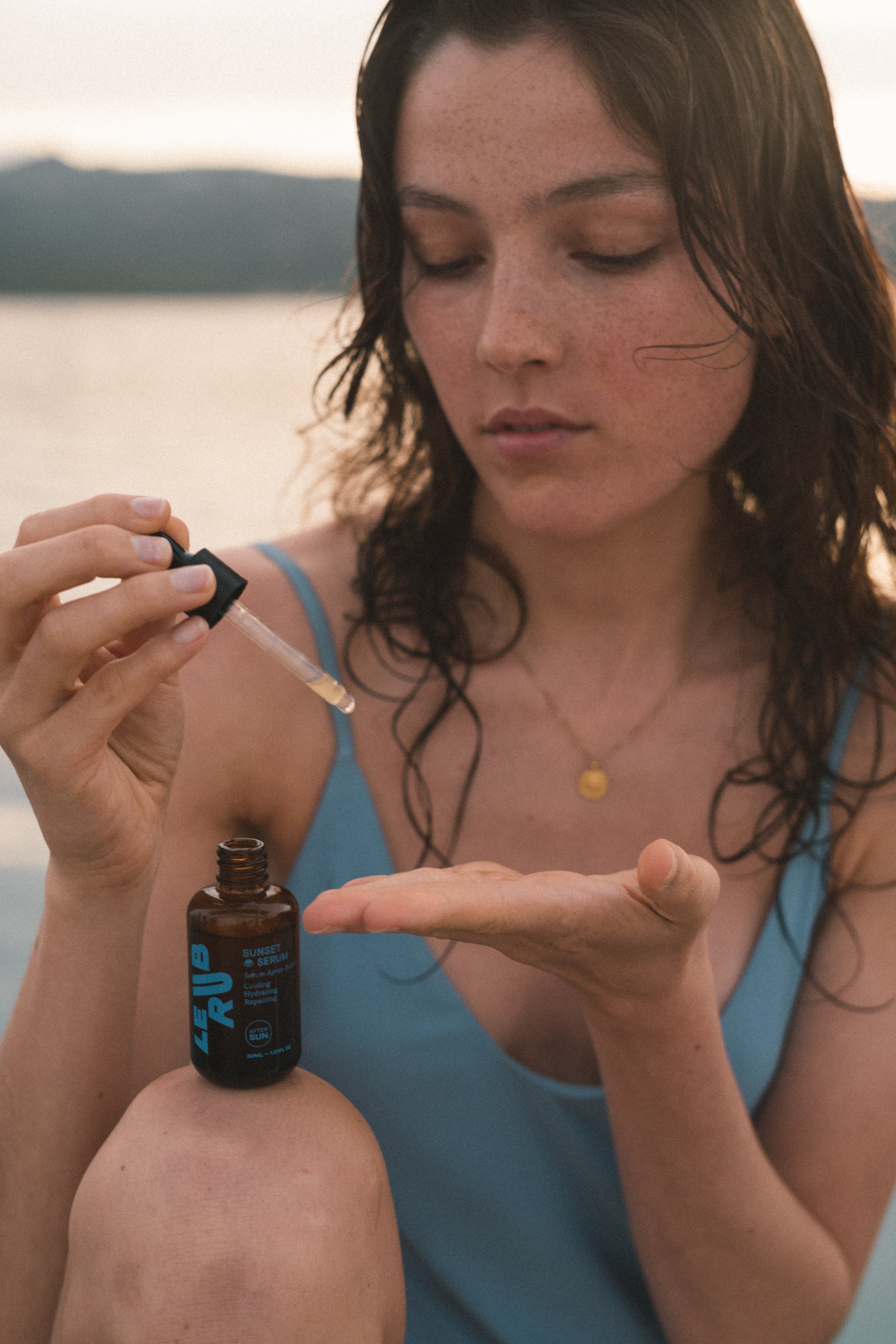 Le Rub Sunset Serum being carefully applied by a woman with wet hair at golden hour, near a water landscape, showcasing natural skincare ritual