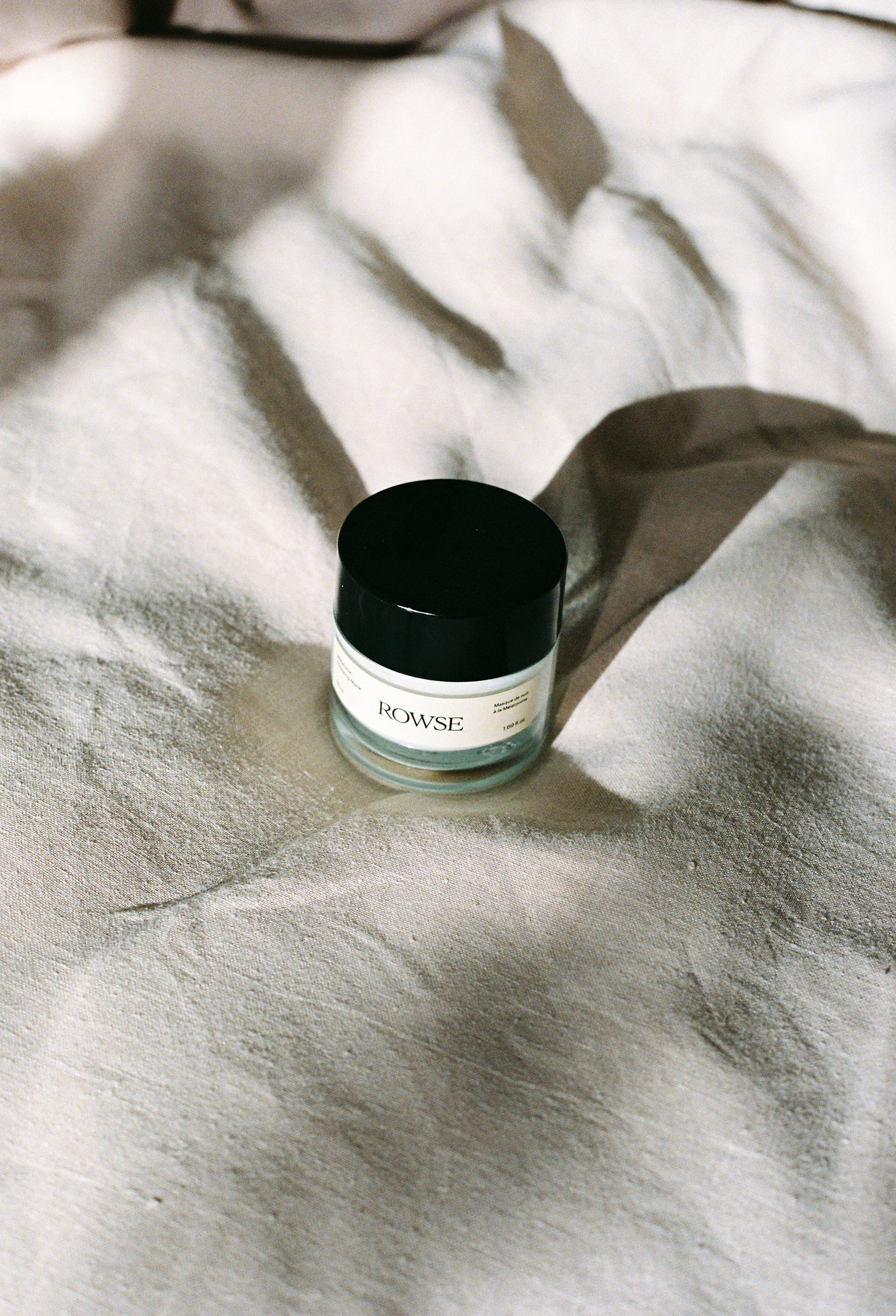 ROWSE Melatonin Sleeping Mask in minimalist glass jar with black lid, positioned on soft textured fabric, showcasing clean skincare design