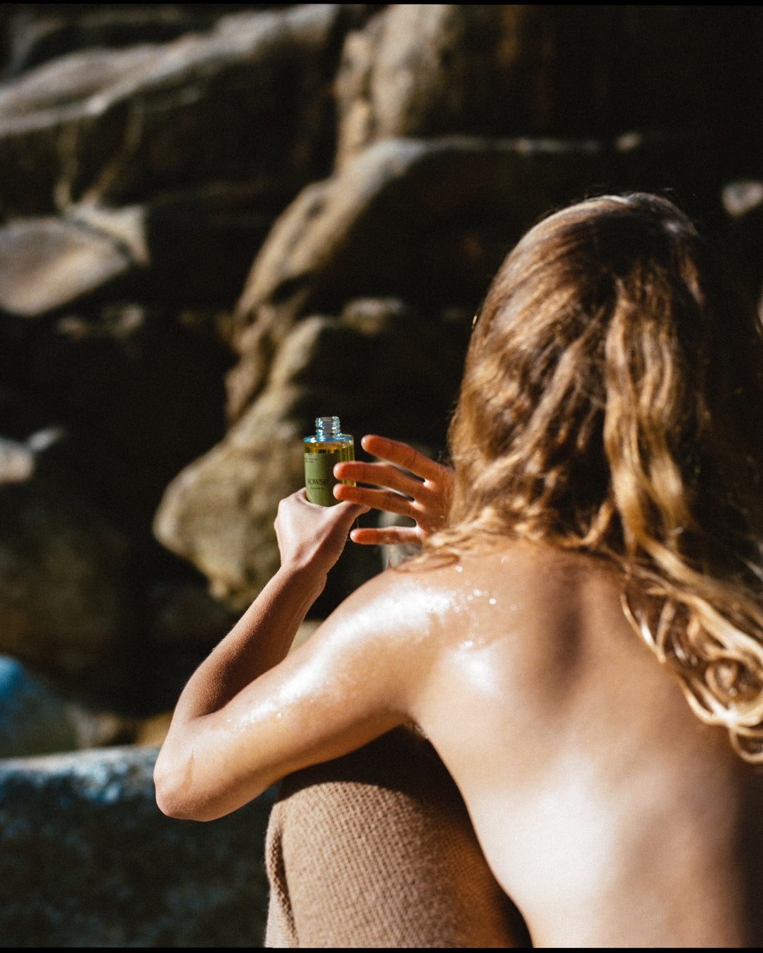ROWSE Winter Body Oil being held by a model with sun-kissed skin, positioned against a natural rocky backdrop, showcasing skincare in an editorial setting