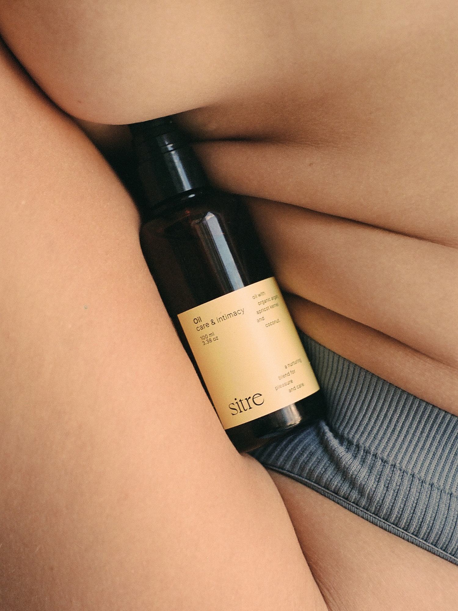 Sitre Oil Care & Intimacy Oil bottle with yellow label positioned between skin and fabric, showcasing intimate wellness product in minimalist, editorial styling