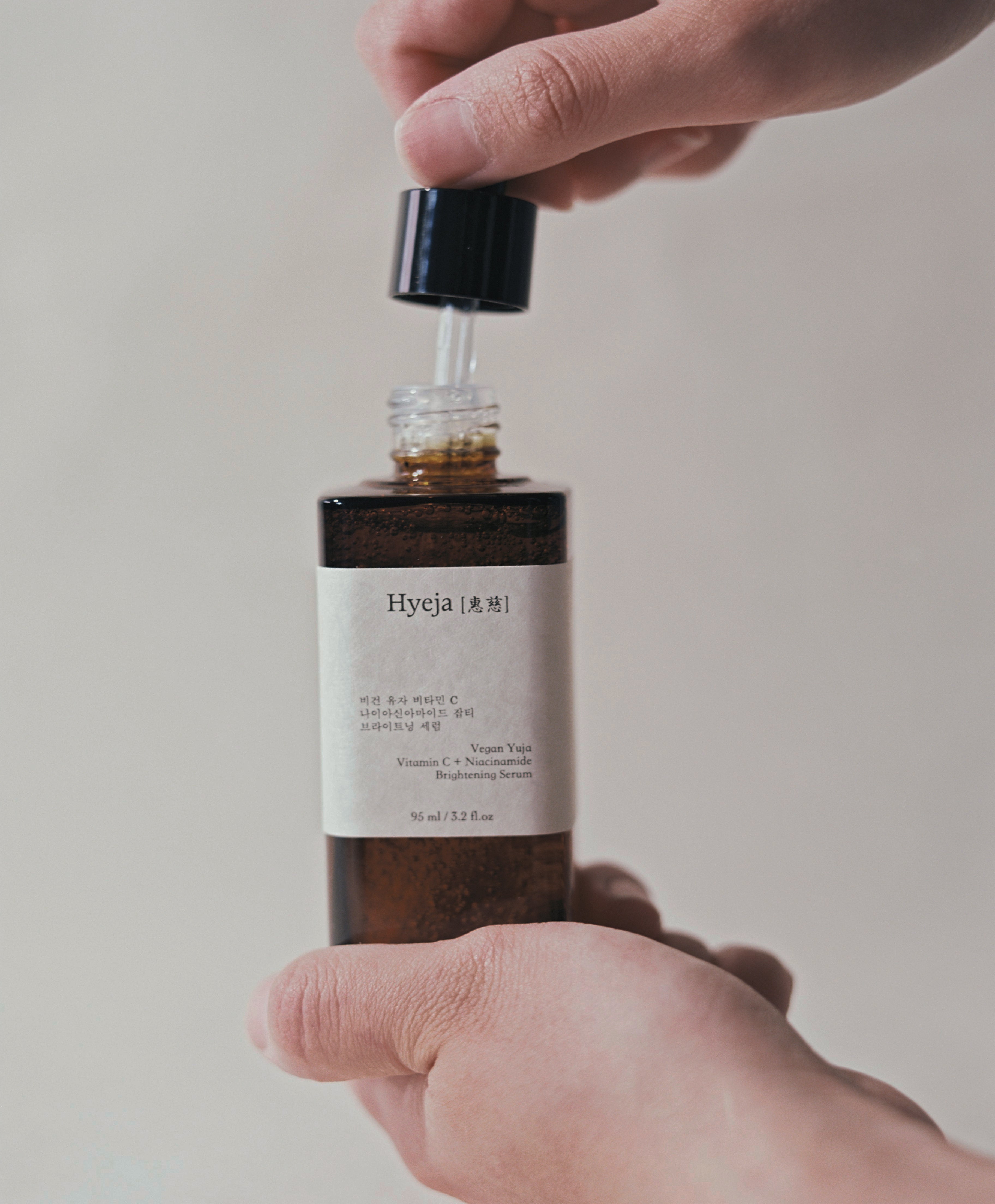 Hyeja Vegan Yuzu Vitamin C + Niacinamide Brightening Serum being dispensed from dropper bottle, showcasing amber-colored formula and minimalist Korean skincare design