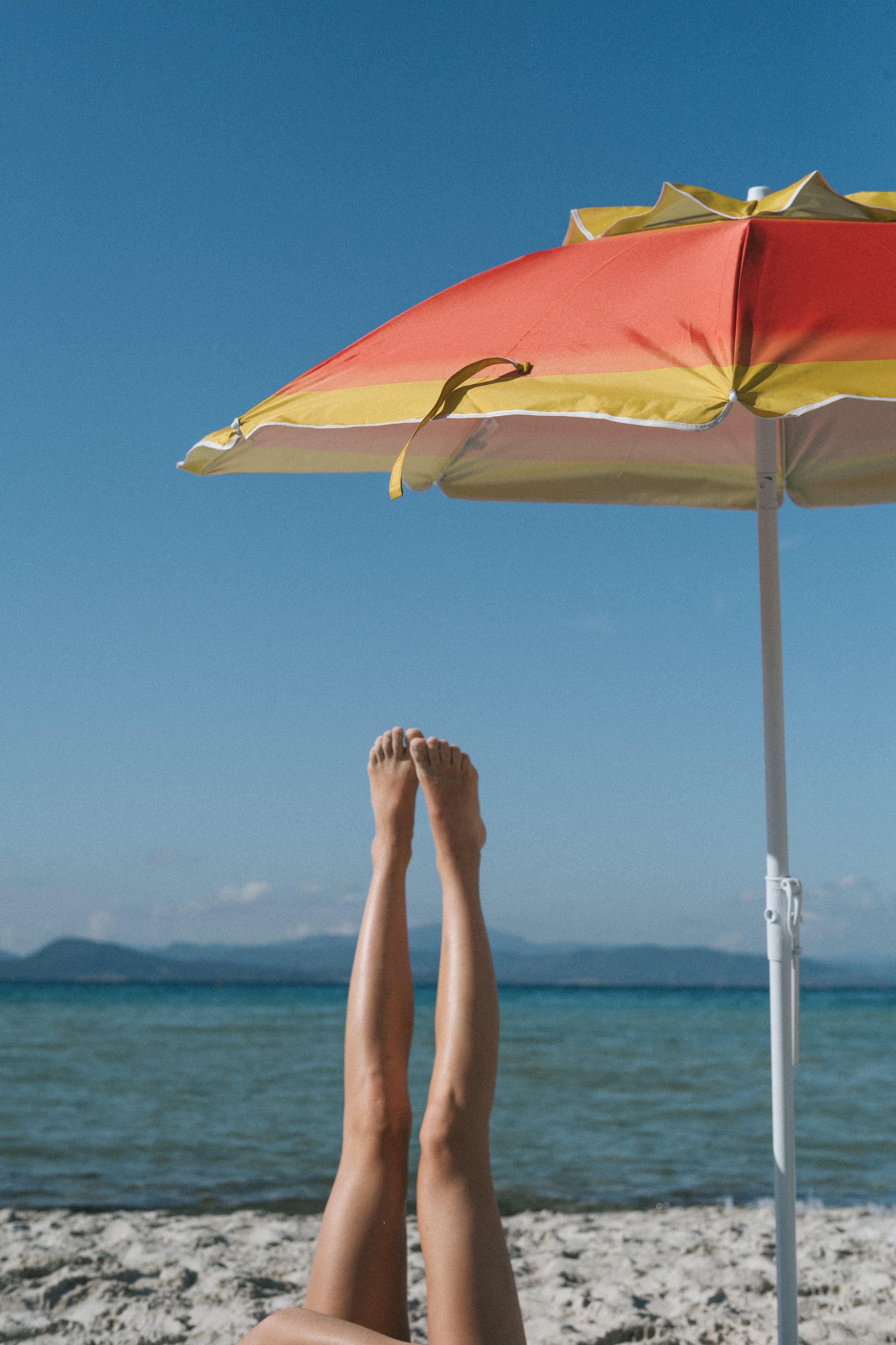 Raised legs under beach umbrella with ocean and mountain backdrop, showcasing Le Rub suncare lifestyle with minimalist, relaxed aesthetic