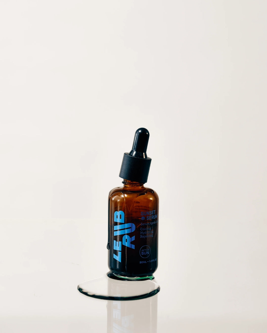 Le Rub After Sun Sunset Serum in amber glass dropper bottle, showcasing minimalist design with blue branding, positioned on reflective surface with neutral background