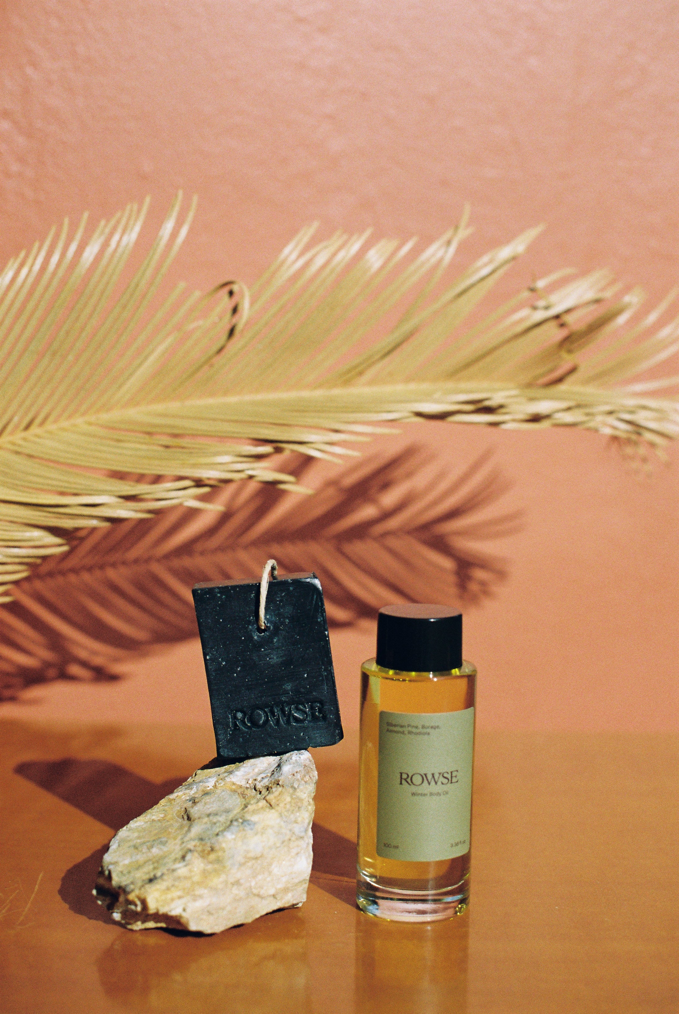 ROWSE Winter Body Oil positioned with a black soap bar and natural stone, set against a warm terracotta background with palm leaf shadows

