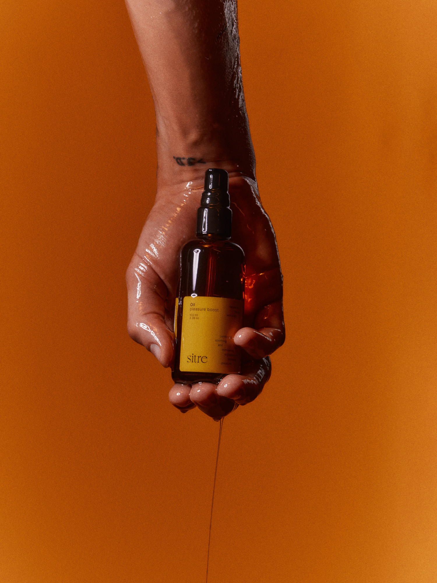 Sitre Oil Care Intimacy Oil bottle with yellow label, held in a glistening hand against a warm orange background, showcasing intimate wellness product
