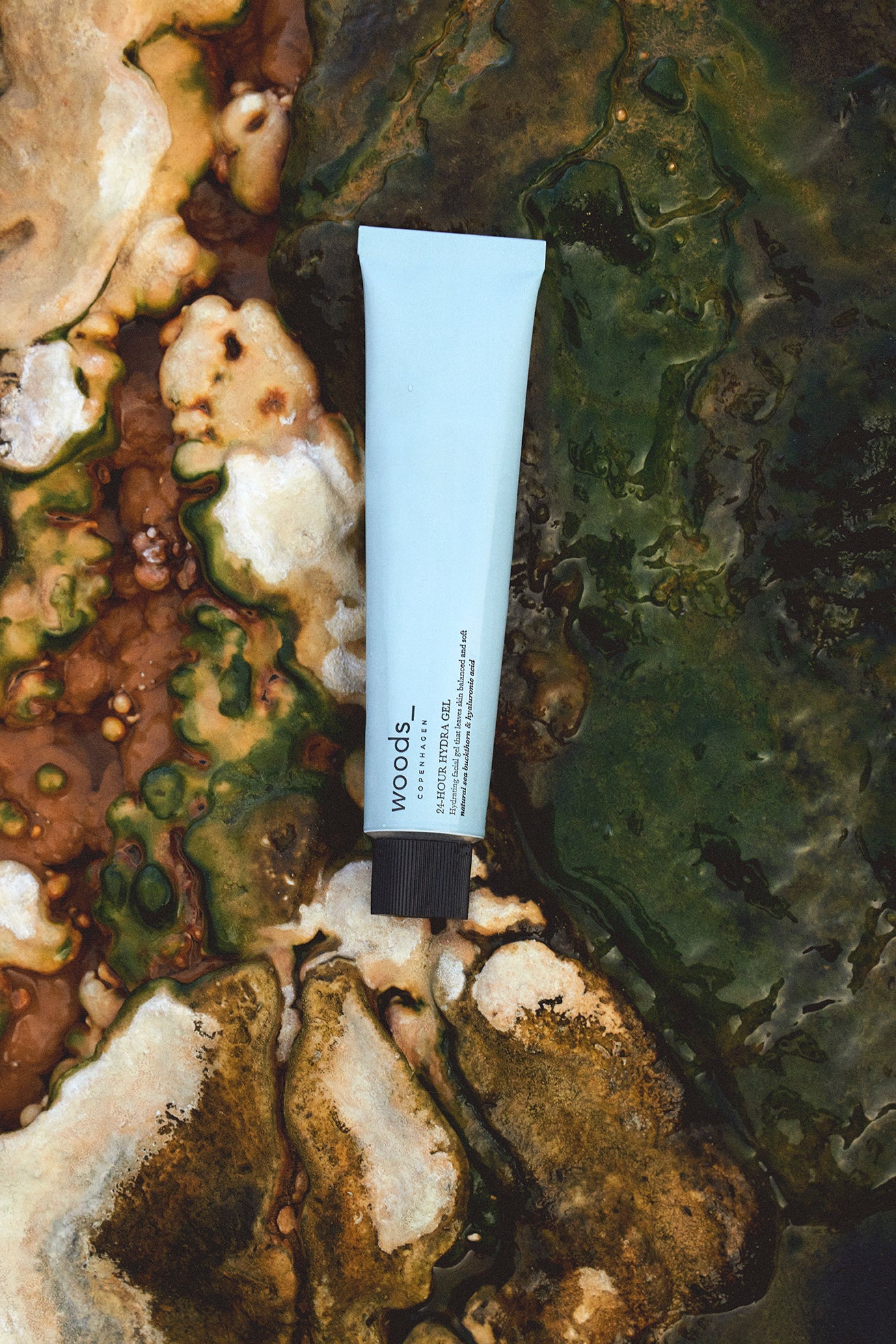 Woods Copenhagen 24-Hour Hydra Gel tube in soft blue, positioned on an organic, textured background with earthy green and brown tones